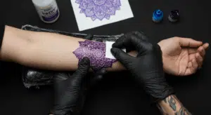 Mãos de tatuador aplicando um stencil de tatuagem roxo detalhado no antebraço de um cliente, com materiais de stencil ao fundo.