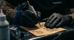 Mãos de tatuador praticando com máquina de tatuar em uma pele artificial, destacando a necessidade de treino para desenvolver a técnica.