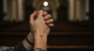 Mãos em oração com uma tatuagem de cruz no pulso, ilustrando a discussão sobre se fazer tatuagem é pecado.