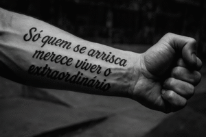 Tatuagem no antebraço com a frase "Só quem se arrisca merece viver o extraordinário" em lettering.