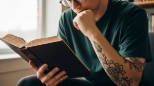 Pessoa com braços tatuados lendo a Bíblia em um momento de estudo, refletindo sobre fé e tatuagem.